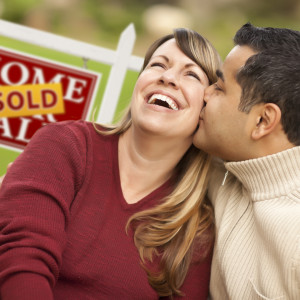 Happy Mixed Race Couple in Front of Sold Real Estate Sign.